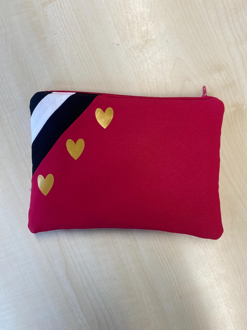 May include: A rectangular red zippered pouch with three gold heart accents. The upper left corner features a black and white striped design. The zipper is red. This pouch is suitable for carrying small items.