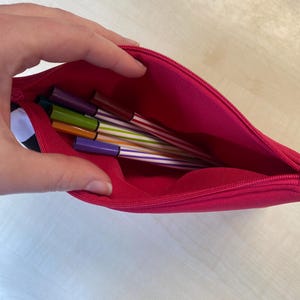 May include: A bright pink pencil case, partially open, revealing several colorful pens. The pens have striped designs in various colors, including purple, green, and orange. The case is made of a smooth fabric and has a zipper closure.