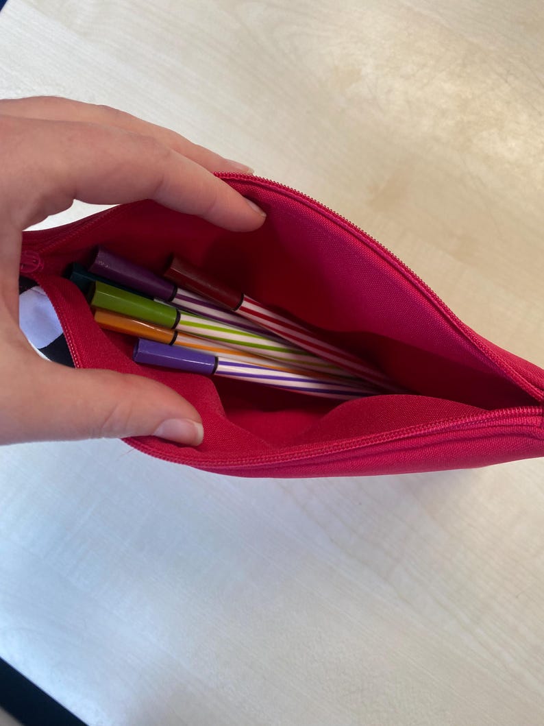 May include: A bright pink pencil case, partially open, revealing several colorful pens. The pens have striped designs in various colors, including purple, green, and orange. The case is made of a smooth fabric and has a zipper closure.