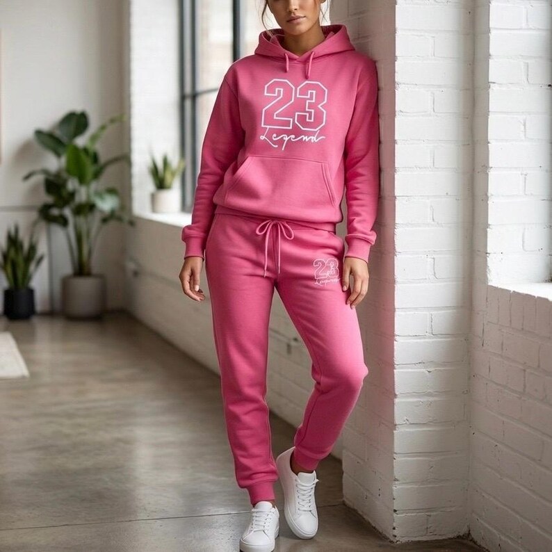 Puede incluir: Un ch&aacute;ndal rosa de dos piezas. La sudadera con capucha presenta el n&uacute;mero "23" y la palabra "Legend" en blanco. Los pantalones de ch&aacute;ndal a juego tienen cintura con cord&oacute;n y el n&uacute;mero "23" impreso en la pierna izquierda. Zapatillas blancas completan el conjunto.
