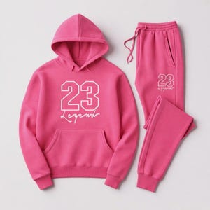 Puede incluir: Un conjunto de ch&aacute;ndal rosa que consta de una sudadera con capucha y un pantal&oacute;n de ch&aacute;ndal. La sudadera con capucha tiene un bolsillo delantero y el n&uacute;mero "23" con la palabra "Legend" en blanco. El pantal&oacute;n de ch&aacute;ndal tiene cintura con cord&oacute;n y el mismo dise&ntilde;o "23 Legend".