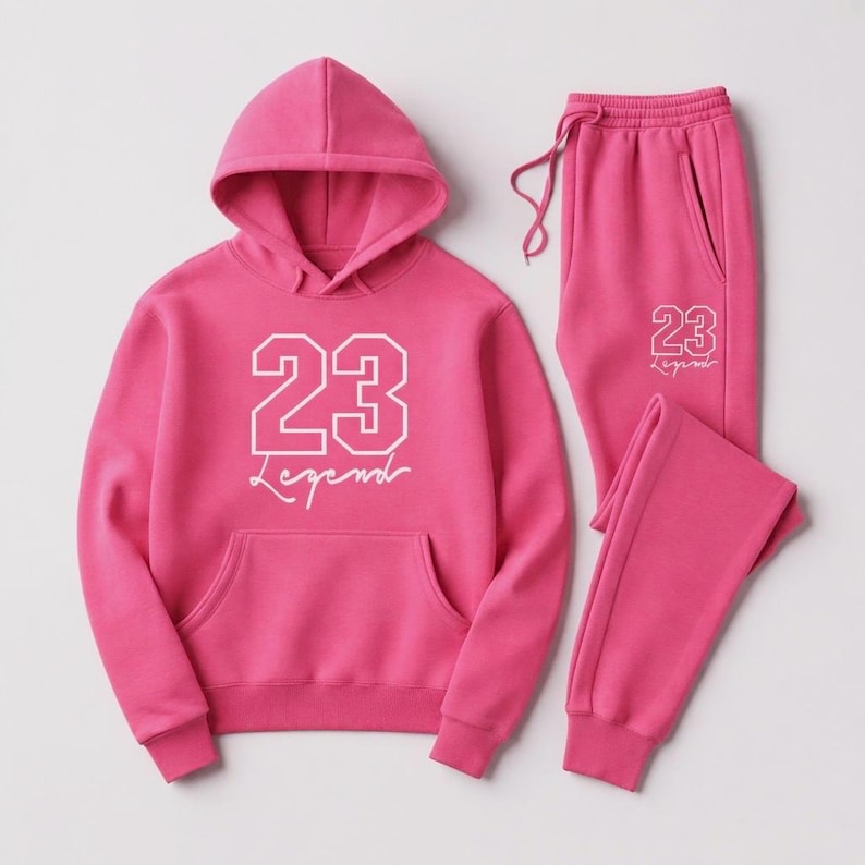 Puede incluir: Un conjunto de ch&aacute;ndal rosa que consta de una sudadera con capucha y un pantal&oacute;n de ch&aacute;ndal. La sudadera con capucha tiene un bolsillo delantero y el n&uacute;mero "23" con la palabra "Legend" en blanco. El pantal&oacute;n de ch&aacute;ndal tiene cintura con cord&oacute;n y el mismo dise&ntilde;o "23 Legend".