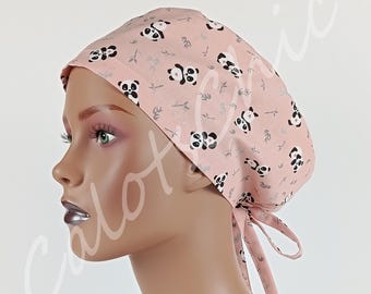 May include: A light pink scrub cap featuring a repeating pattern of black and white panda bears and small green bamboo stalks. The cap has a rounded crown and includes two fabric ties at the back for an adjustable fit, shown on a mannequin head.