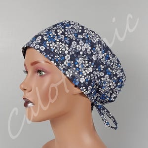Op de afbeelding: Een operatiemuts met een donkerblauwe achtergrond en een dicht patroon van kleine witte en lichtblauwe bloemen. De muts heeft een ronde bovenkant, een brede voorband en twee stoffen linten aan de achterkant voor een goede pasvorm.