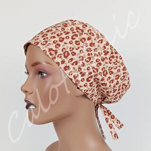 Puede incluir: Un gorro quirúrgico con fondo beige y un patrón de leopardo marrón repetitivo. El gorro tiene una forma redondeada y dos cintas de tela en la parte posterior para ajustar, proporcionando un ajuste seguro con un diseño clásico de lazo.