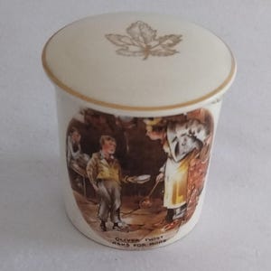 Könnte beinhalten: Eine cremefarbene Keramikdose mit Deckel, Goldrand und Blattdesign. Der Korpus zeigt eine Szene aus "Oliver Twist" mit dem Text "Oliver Twist Asks For More".