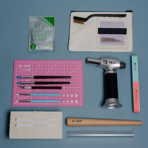 May include: A metal clay crafting kit including a silver clay packet, a canvas pouch with a wire brush and blade, a pink A5 cutting mat, three brushes, two stylus tools, a butane torch, a nail buffer, a ceramic block, a ring mandrel, and a tube.