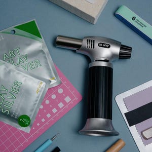 May include: A silver and black butane torch with the text OK OKAY, two silver packets of Art Clay Silver, a pink grid mat, a light blue nail buffer, a white stone block, a metal ruler, and various small crafting tools on a blue surface.