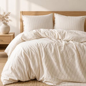 May include: Cream-colored bedding set with a subtle striped pattern. The set includes a duvet cover and two pillowcases. The pillows are against a light wood headboard. The overall aesthetic is simple and inviting, perfect for a bedroom.