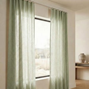 Puede incluir: Dos cortinas a rayas verdes y blancas cuelgan de una barra plateada frente a una ventana. Las cortinas son de tela ligera y ligeramente translúcidas. La habitación tiene paredes claras y suelo de madera.