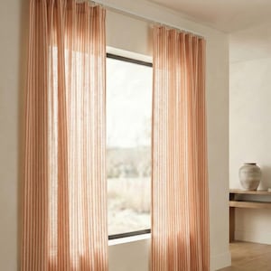 May include: Peach-coloured striped curtains hanging in front of a window. The curtains are made of a light, semi-sheer fabric with vertical stripes. The room has a neutral colour palette with a wooden floor.