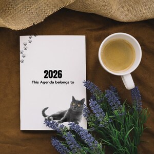 May include: A white agenda book for 2026 with a gray cat illustration, paw prints, and the text "This Agenda belongs to." A white coffee cup and lavender sprigs are also present on a brown surface.