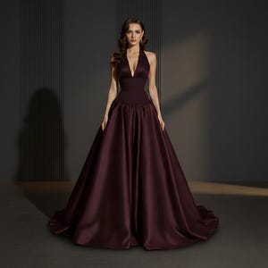 May include: A floor-length, deep burgundy gown featuring a sleeveless halter neckline with a plunging V-cut bodice. The dress has a fitted waistline that transitions into a full, voluminous skirt with a smooth, satin-like finish and a train.