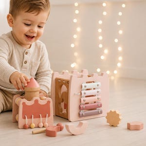 May include: A wooden activity cube toy featuring a castle design in shades of pink and natural wood. The toy includes a built-in xylophone with metal bars, bead maze elements, and various geometric wooden shapes for sorting and play activities.