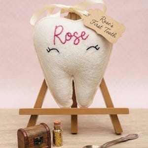 Può includere: Un cuscino in feltro bianco a forma di dente con il nome Rose ricamato in rosa e occhi cuciti in nero. È appeso a un cavalletto di legno con un'etichetta beige che riporta la scritta Rose's First Tooth. Sotto si trovano un piccolo forziere in legno, una boccetta di vetro con glitter dorati e un cucchiaio d'argento che contiene un singolo dente.