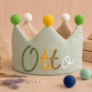 Può includere: Una corona in tessuto verde chiaro con il nome "Otto" ricamato in verde, giallo e bianco. La corona è sormontata da pompon verdi, bianchi e gialli. Altri oggetti includono forbici, lavanda e palline decorative.