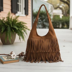 May include: A brown suede shoulder bag with long fringe detailing. The bag has a curved top and a long strap. The fringe is layered and hangs down from the bottom of the bag. The bag is sitting on a white wooden surface.