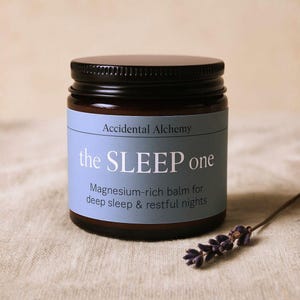 May include: A brown glass jar with a black lid and a light blue label. The label says "Accidental Alchemy the SLEEP one" and "Magnesium-rich balm for deep sleep & restful nights." A sprig of lavender is next to the jar.
