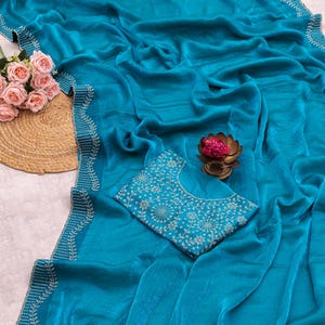 May include: A teal saree with intricate white embroidery along the borders. The saree is draped, showcasing its flowing fabric. A matching teal blouse with detailed white floral embroidery is also present. A bouquet of pink roses and a decorative lotus flower are also visible.