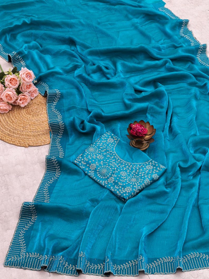 May include: A teal saree with intricate white embroidery along the borders. The saree is draped, showcasing its flowing fabric. A matching teal blouse with detailed white floral embroidery is also present. A bouquet of pink roses and a decorative lotus flower are also visible.