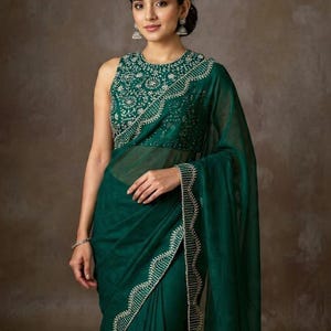 May include: A dark emerald green saree with intricate silver embroidery along the borders and neckline. The sleeveless blouse features detailed silver embellishments. The saree is draped elegantly, showcasing the flowing fabric and design.