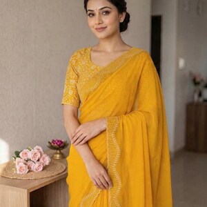 May include: A woman wearing a bright yellow saree with intricate embroidered detailing along the border and on the short-sleeved blouse. The saree is draped elegantly, and the woman is smiling. A small table with pink roses and a decorative item is in the background.