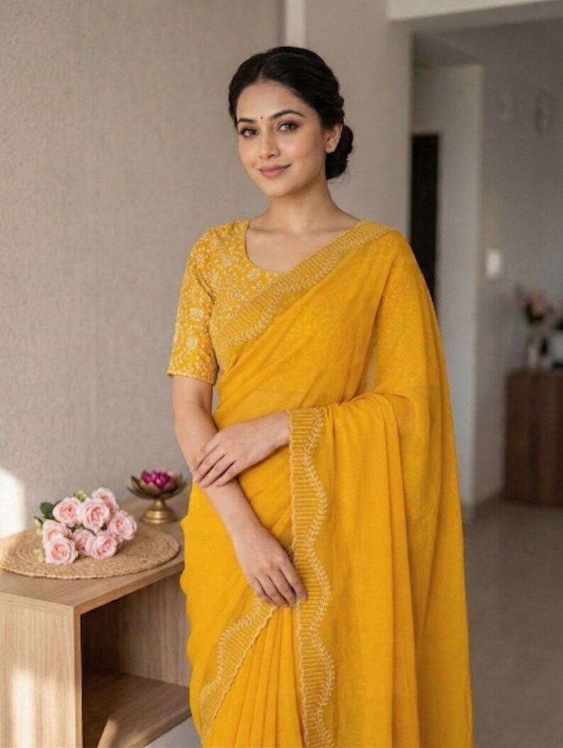 May include: A woman wearing a bright yellow saree with intricate embroidered detailing along the border and on the short-sleeved blouse. The saree is draped elegantly, and the woman is smiling. A small table with pink roses and a decorative item is in the background.