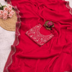 May include: A vibrant red saree with intricate gold-colored embroidery along the edges and a matching blouse. The saree is draped, showcasing its flowing fabric and detailed design. The blouse features a similar floral pattern.