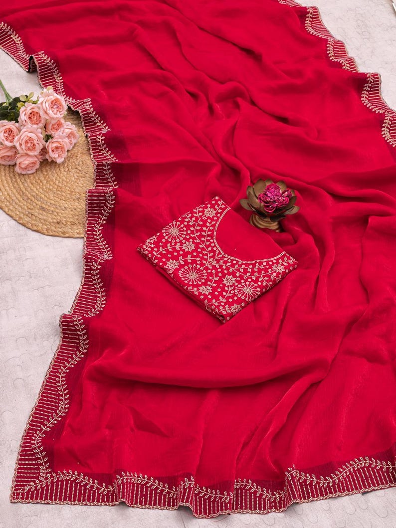 May include: A vibrant red saree with intricate gold-colored embroidery along the edges and a matching blouse. The saree is draped, showcasing its flowing fabric and detailed design. The blouse features a similar floral pattern.