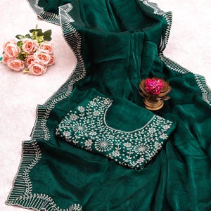 May include: A dark emerald green saree with intricate white embroidery along the borders and on the matching blouse. The saree is draped, showcasing its flowing fabric and detailed design. A small bouquet of pink roses and a decorative flower are also present.