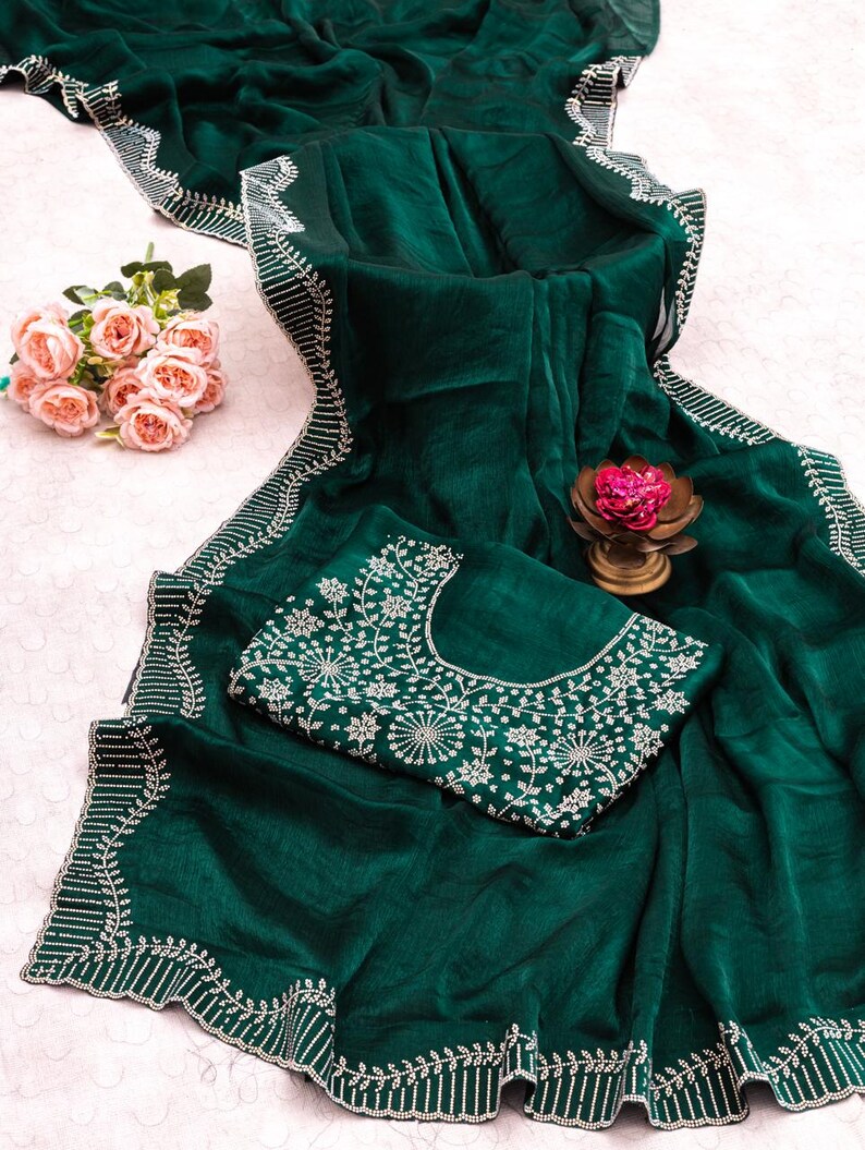 May include: A dark emerald green saree with intricate white embroidery along the borders and on the matching blouse. The saree is draped, showcasing its flowing fabric and detailed design. A small bouquet of pink roses and a decorative flower are also present.