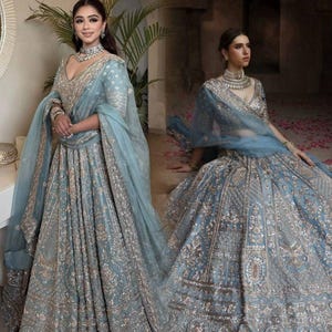May include: A dusty blue lehenga set featuring a V-neck blouse and a voluminous floor-length skirt, both heavily embellished with intricate silver and gold metallic embroidery. A matching sheer dupatta is draped over the shoulder.
