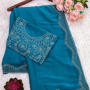 May include: Teal-colored saree with intricate white embroidery along the borders and on the blouse. The saree is draped over a wooden hanger, with a bouquet of pink roses and a decorative flower adding a touch of elegance.