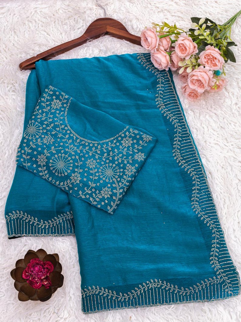 May include: Teal-colored saree with intricate white embroidery along the borders and on the blouse. The saree is draped over a wooden hanger, with a bouquet of pink roses and a decorative flower adding a touch of elegance.
