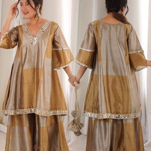 May include: A two-piece, gold and beige outfit with a V-neck top and matching skirt. The top has a mirrored trim along the neckline and sleeves. The skirt is a wide-leg style. The outfit is made of a shiny, flowing fabric.