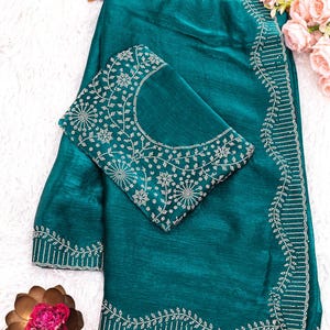 May include: Teal-colored saree with intricate white embroidery along the borders and on the blouse. The blouse features a detailed floral pattern around the neckline. The saree is draped over a wooden hanger, with a decorative flower arrangement in the background.