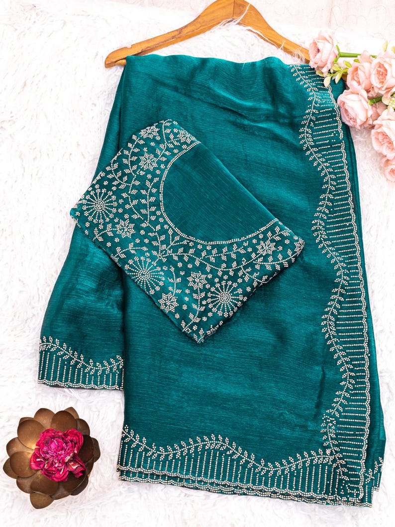 May include: Teal-colored saree with intricate white embroidery along the borders and on the blouse. The blouse features a detailed floral pattern around the neckline. The saree is draped over a wooden hanger, with a decorative flower arrangement in the background.
