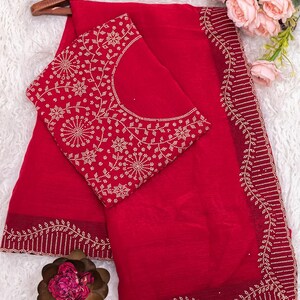 May include: A vibrant red saree set with intricate gold-colored embroidery. The saree features a detailed border design, and the matching blouse has a floral pattern. The set is displayed on a wooden hanger, with a soft, white background.