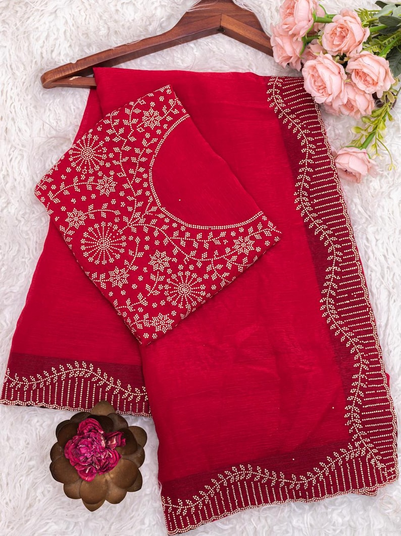 May include: A vibrant red saree set with intricate gold-colored embroidery. The saree features a detailed border design, and the matching blouse has a floral pattern. The set is displayed on a wooden hanger, with a soft, white background.