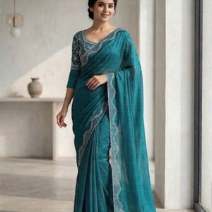 May include: A teal saree with a detailed silver border and matching three-quarter sleeve blouse. The blouse features a patterned design on the sleeves and neckline. The saree is draped elegantly, showcasing its flowing fabric and intricate details.