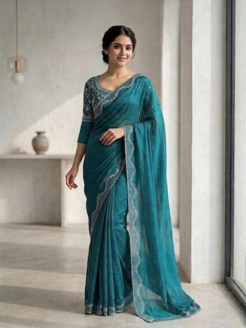 May include: A teal saree with a detailed silver border and matching three-quarter sleeve blouse. The blouse features a patterned design on the sleeves and neckline. The saree is draped elegantly, showcasing its flowing fabric and intricate details.