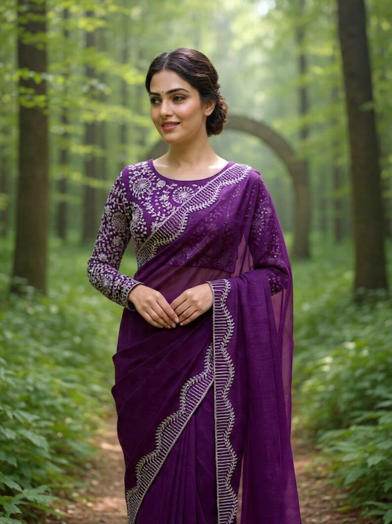 May include: A woman wearing a deep purple saree with intricate silver embroidery. The long-sleeved blouse features detailed floral patterns, and the saree's border is embellished with vertical silver lines. The background is a lush green forest.