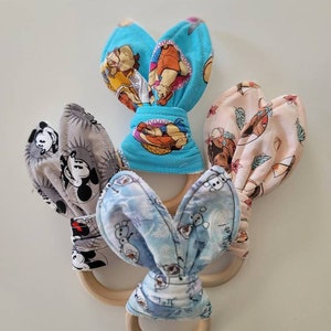 May include: Three wooden teethers with fabric bunny ears. The teethers are made of natural wood and have a variety of colorful fabric patterns, including Mickey Mouse, Frozen, and a tropical print.