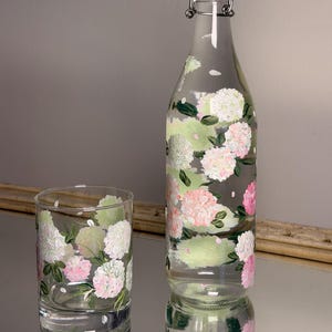 May include: A clear glass bottle with a swing-top stopper and a matching glass tumbler, both hand-painted with clusters of pink and white hydrangea flowers and green leaves. The bottle features a white and red plastic stopper with metal hardware.