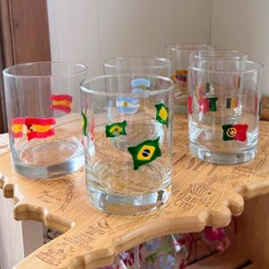 Pode incluir: Um conjunto de cinco copos transparentes, cada um decorado com pequenas bandeiras nacionais pintadas à mão. Os copos estão dispostos sobre uma tábua de madeira clara em forma de mapa, com texto gravado e detalhes geográficos.