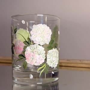 May include: A clear glass tumbler featuring hand-painted floral clusters in shades of white and soft pink. The design includes detailed green leaves and stems, with small scattered petals painted around the flowers on the exterior surface.
