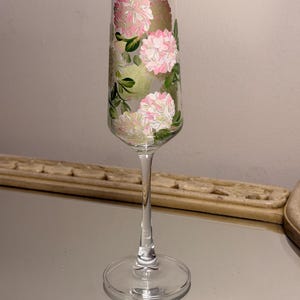 May include: A clear glass champagne flute featuring a hand-painted design of pink and white hydrangea flowers with green leaves. The tall, slender glass has a gold-colored rim at the top and a transparent stem leading to a circular base.