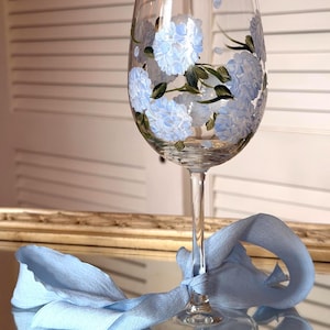 May include: A clear glass wine goblet featuring hand-painted light blue hydrangea flowers with green leaves and stems. A light blue fabric ribbon is tied around the stem of the glass, which rests on a reflective surface.