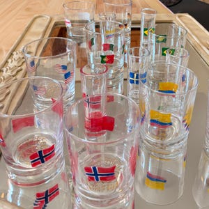 Może przedstawiać: Kolekcja przezroczystych szklanek i kieliszków do alkoholu ustawionych na lustrzanej powierzchni. Każde naczynie ozdobione jest namalowanymi flagami państwowymi, w tym Stanów Zjednoczonych, Norwegii, Portugalii, Brazylii, Szwecji oraz Kolumbii.