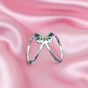 Könnte beinhalten: Ein silberfarbener Ring mit einem einzigartigen Design, bestehend aus zwei gebogenen Bändern, die sich in der Mitte treffen. Der Ring ist mit zwei kleinen, runden, grünen Edelsteinen besetzt. Der Ring befindet sich vor einem rosa Satinhintergrund.